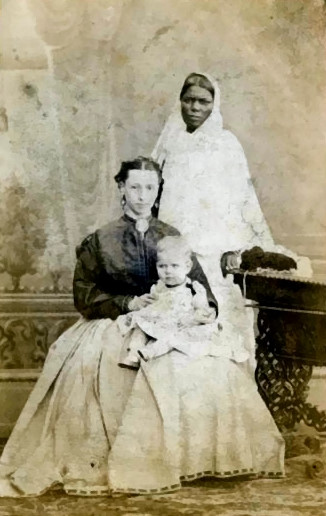 Seated white woman in Victorian clothing with a baby on her lap. Behind her stands an Indian ayah, in traditional Indian clothing.