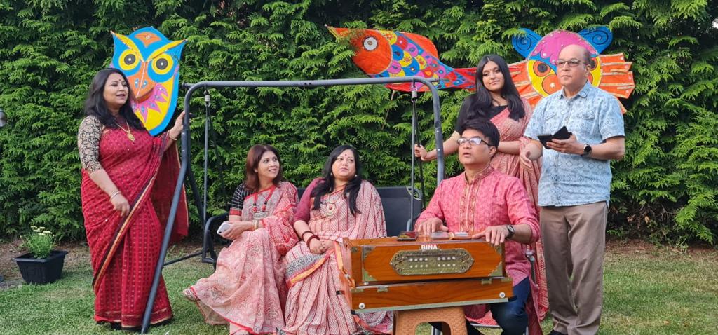 6 members of the Anamika Cultural group standing and sitting outdoors with a wooden musical instrument and brightly colored painted animal-shaped card banners