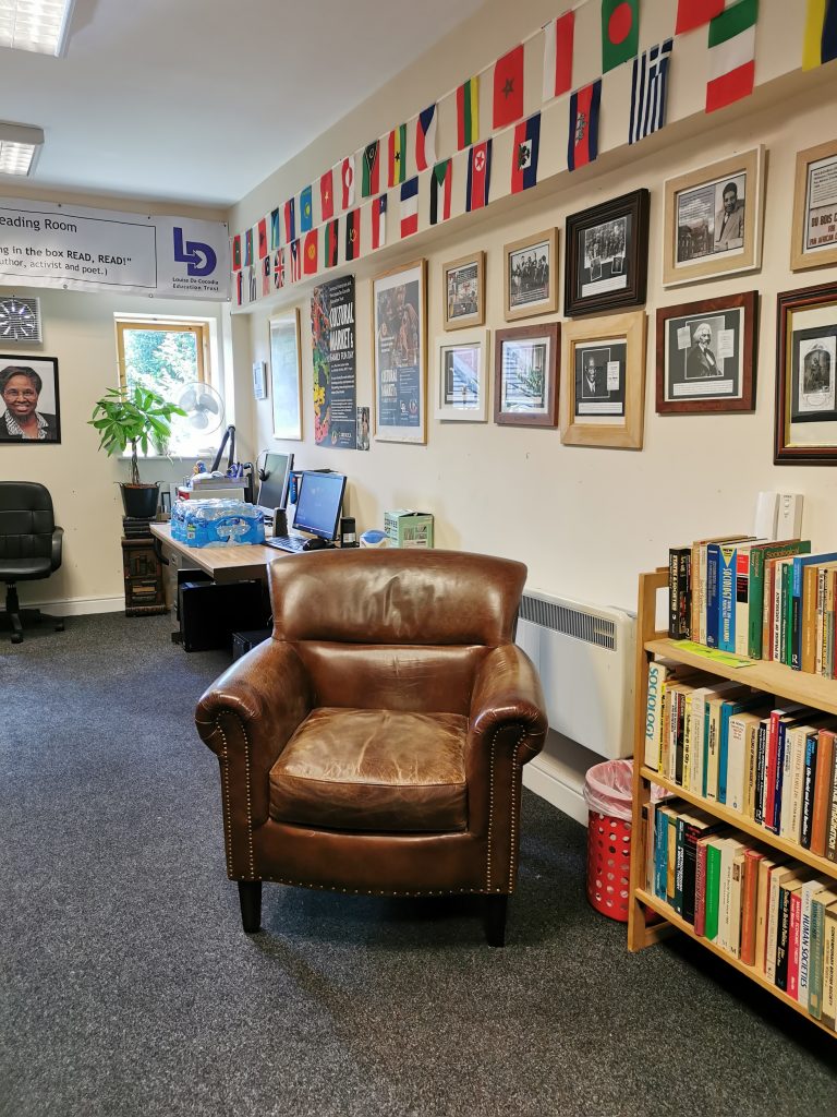 Corner of room. In foreground set of low bookshelves filled with books, padded leather armchair. Walls above displaying various photographs of Black historical figures and posters for Black cultural events, and a variety of national flags. In background desk and window. picture of Louise Da-Cocodia above office chair