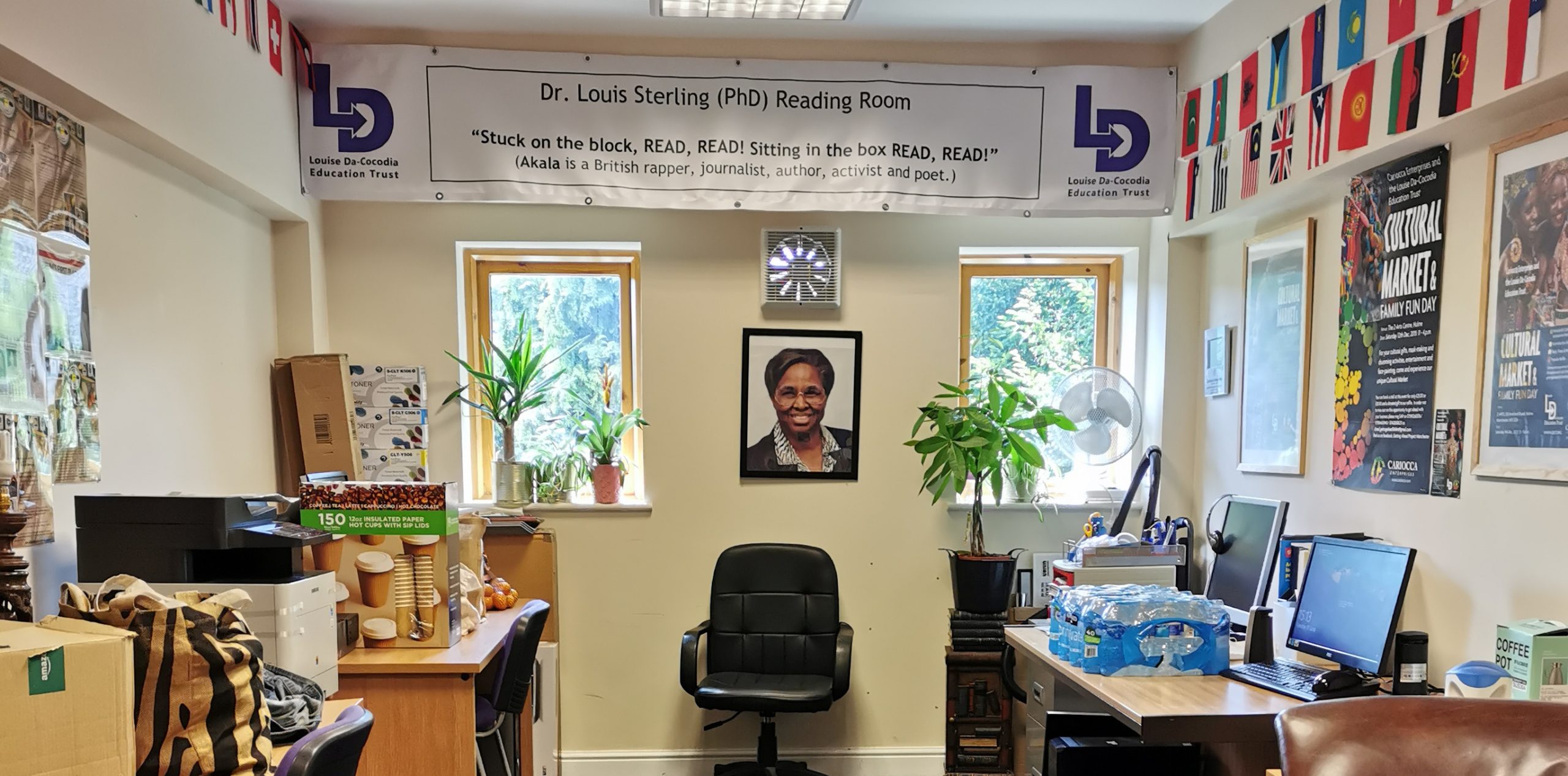 View of office space with desks on either side holding packeets of disposable cups and bottled water. There is a comfortable office chair against the end wall, which has two windows holding plant pots. Between them is a photograph of Louise Da-Cocodia. At the top of the wall is a banner reading Dr Louis Sterling PHD Reading Room "Stuck on the block, read, read. Sitting in the box, read, read." (Akala is a British rapper, journalist, author, activist and poet)