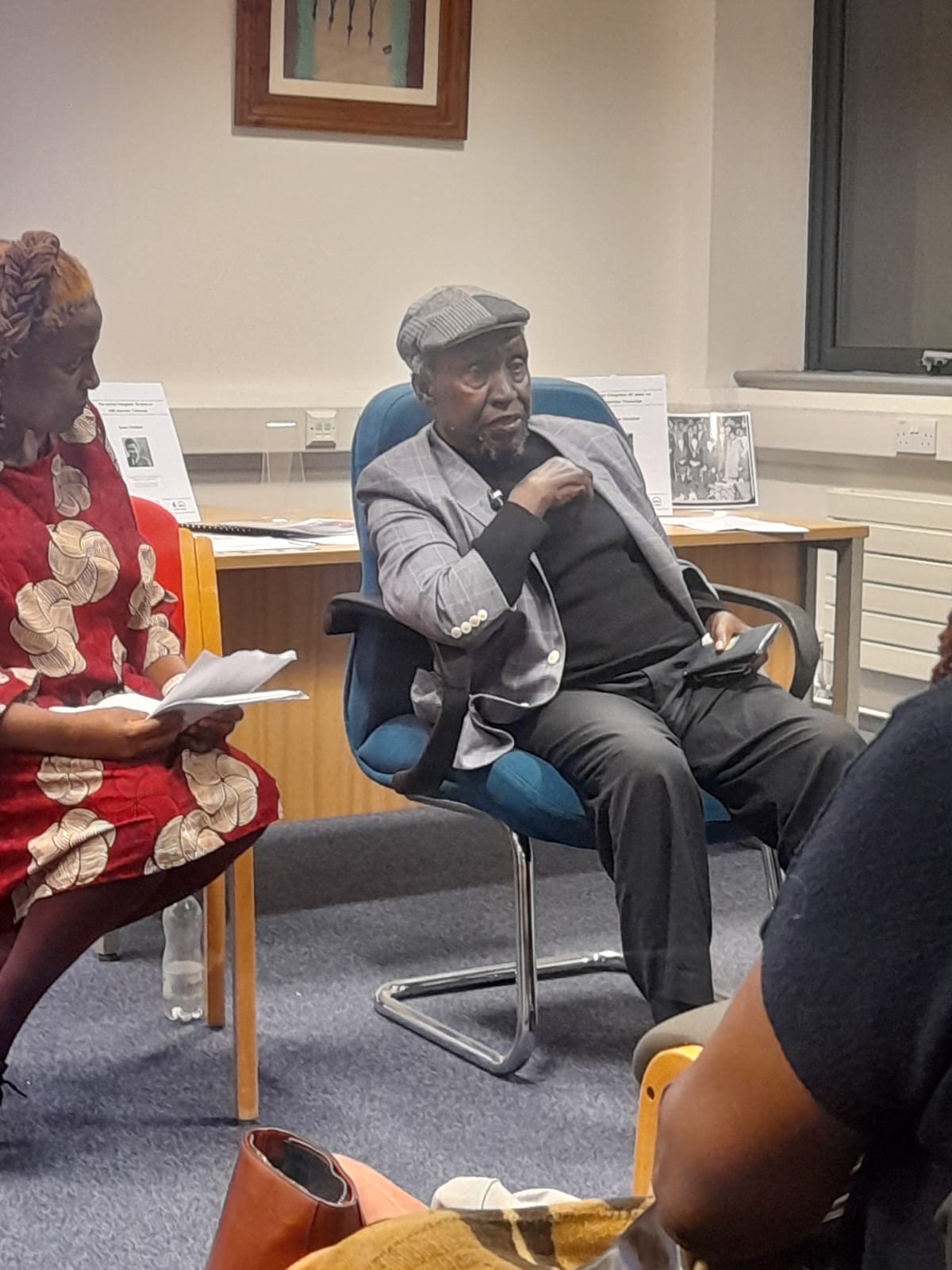 Professor Ngugi Wa Thiong'o sat in a chair facing and talking to the audience. Woman with red dress sits in the chair on the left.